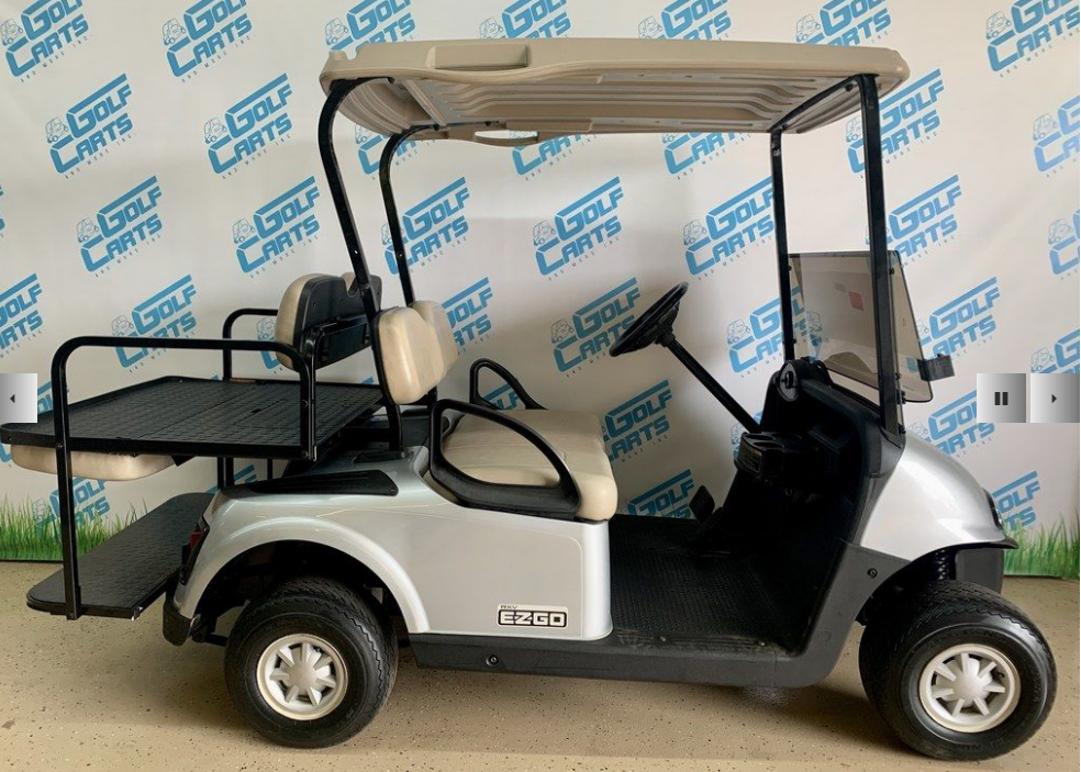 A silver golf cart with rear cargo space, displayed indoors against a branded backdrop.